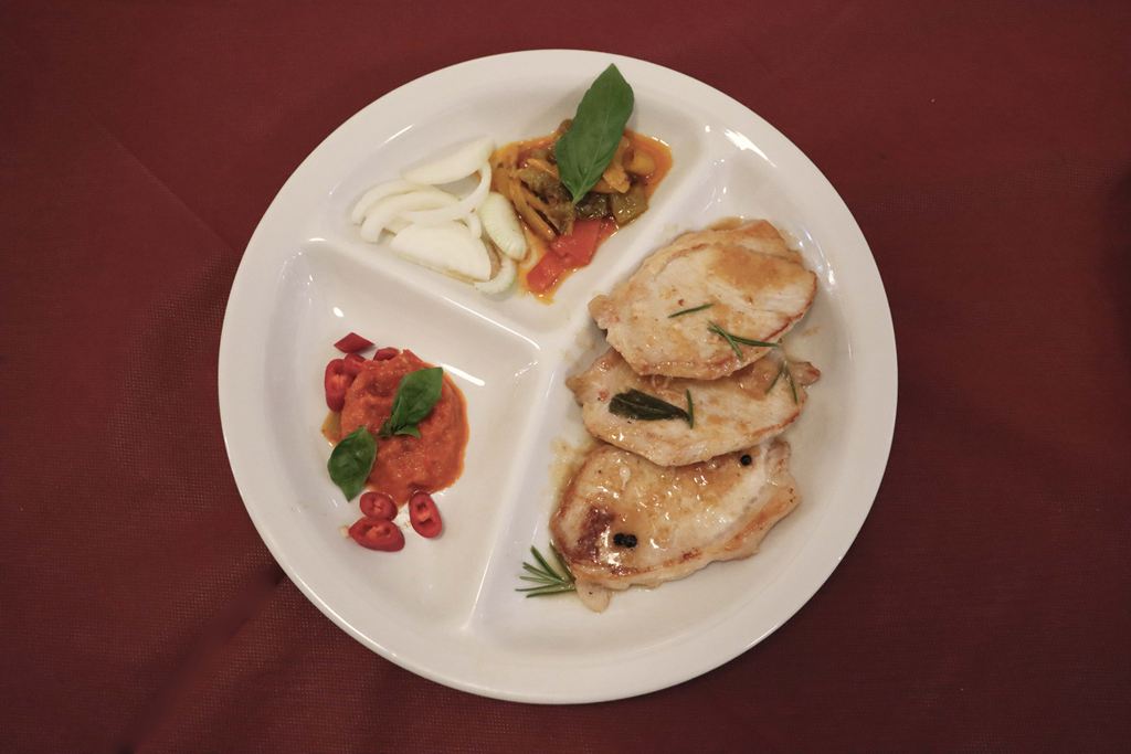 Piatto a scomparti visto dall'alto: fettine di lonza al rosmarino, verdure e una salsa di peperoni con peperoncino