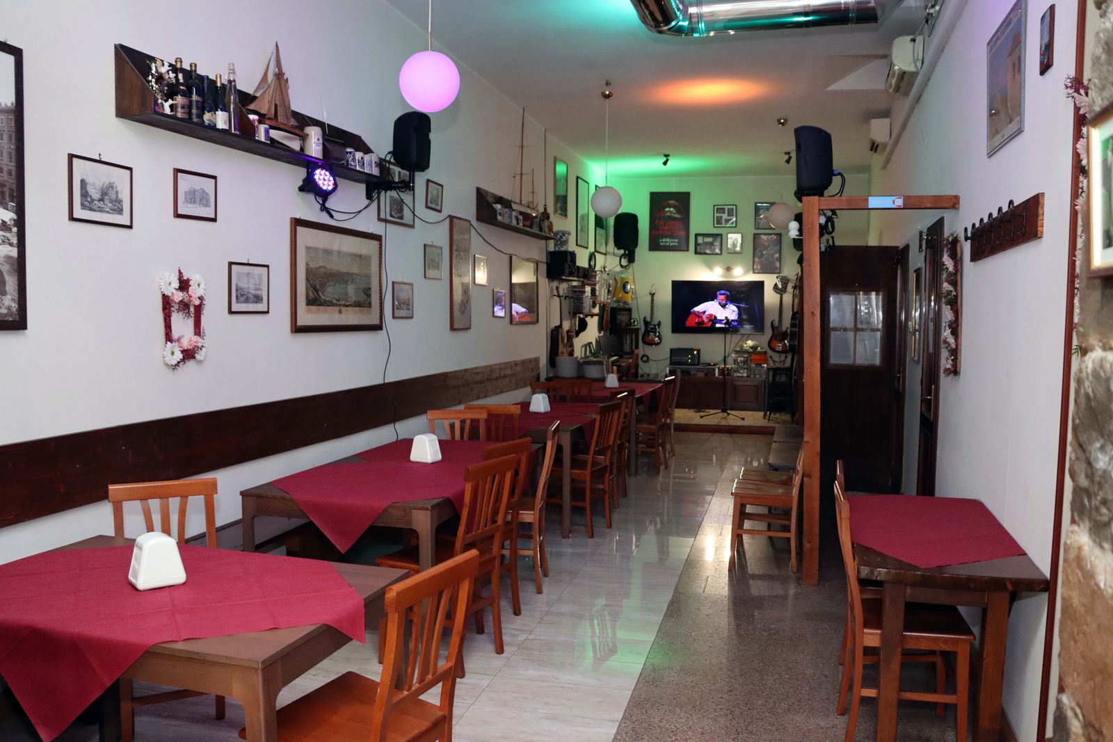 La sala del Buffet da Vittorio: tavoli con le tovaglie bordeaux, sedie in legno, cornici alle pareti e in fondo il piccolo palco con le chitarre
